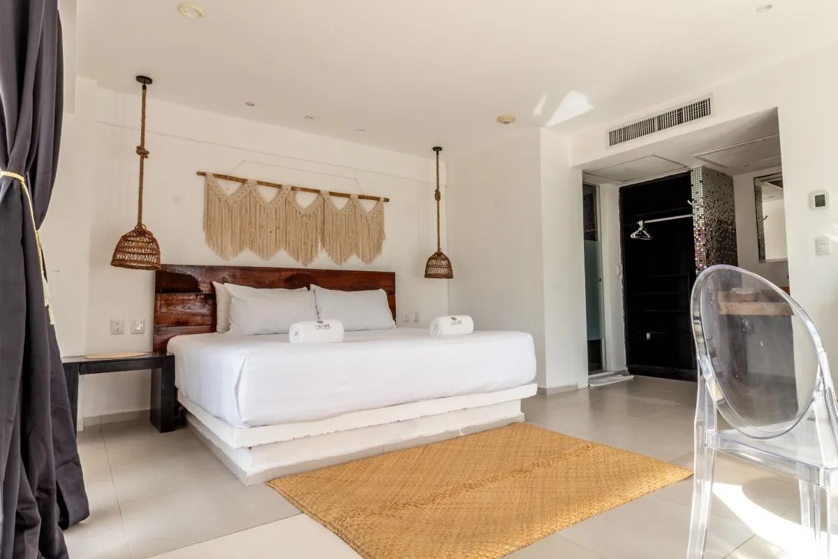 A bright, airy hotel room with a king bed, macramé wall decor, pendant lights, and a clear chair on a woven area rug.