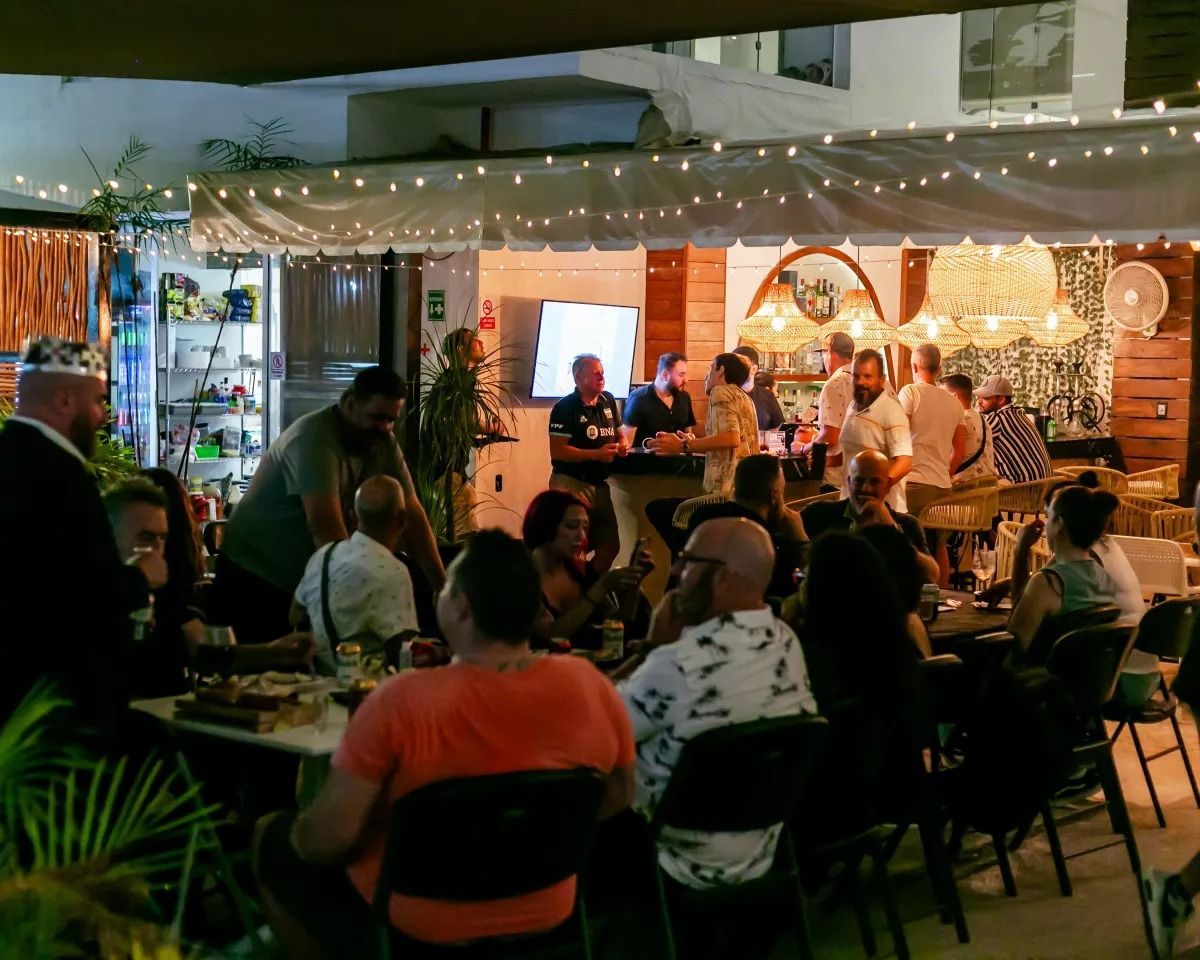 A crowded outdoor patio bar at night, decorated with string lights, filled with people socializing at tables and a bar.