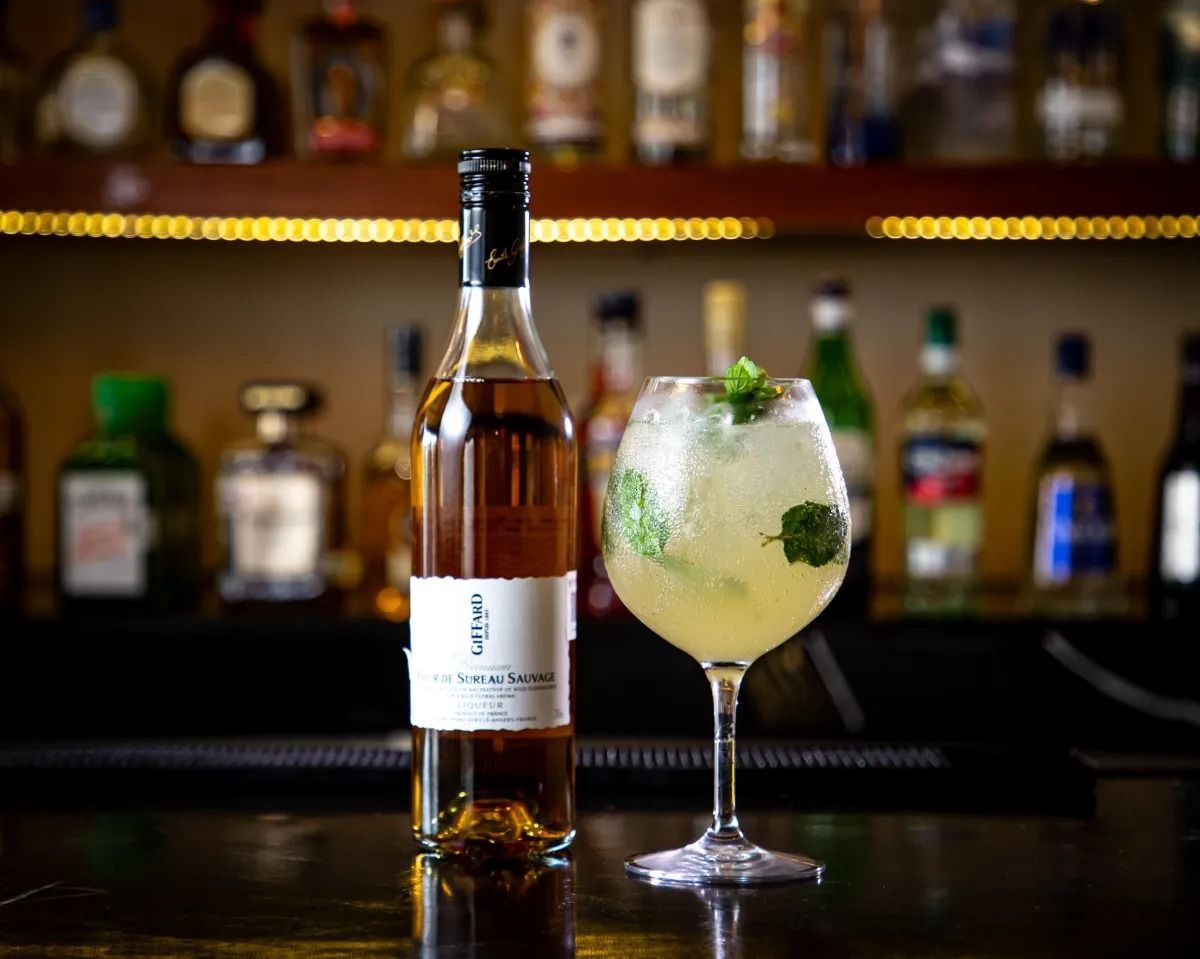 A bottle of amber liqueur and a tall glass of garnished cocktail sitting on a bar counter with blurred bottles behind.