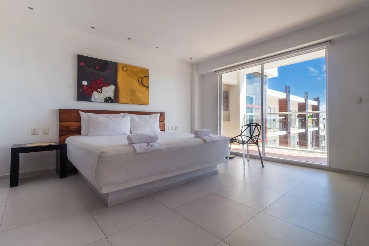 A bright, modern hotel room with a white bed, dark wooden headboard, minimalist black furniture, and a balcony view.