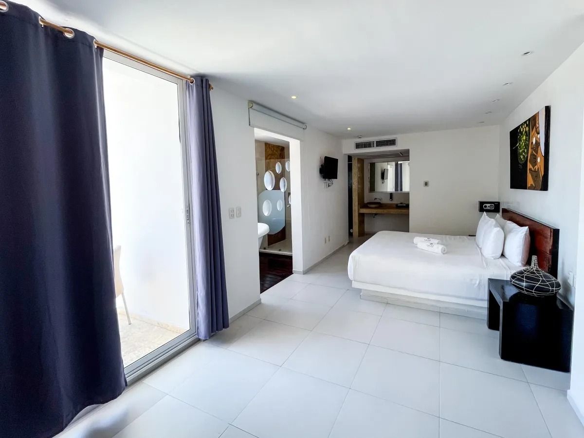 A bright, modern hotel room with a white bed, dark curtains, a balcony view, and an adjacent bathroom doorway.
