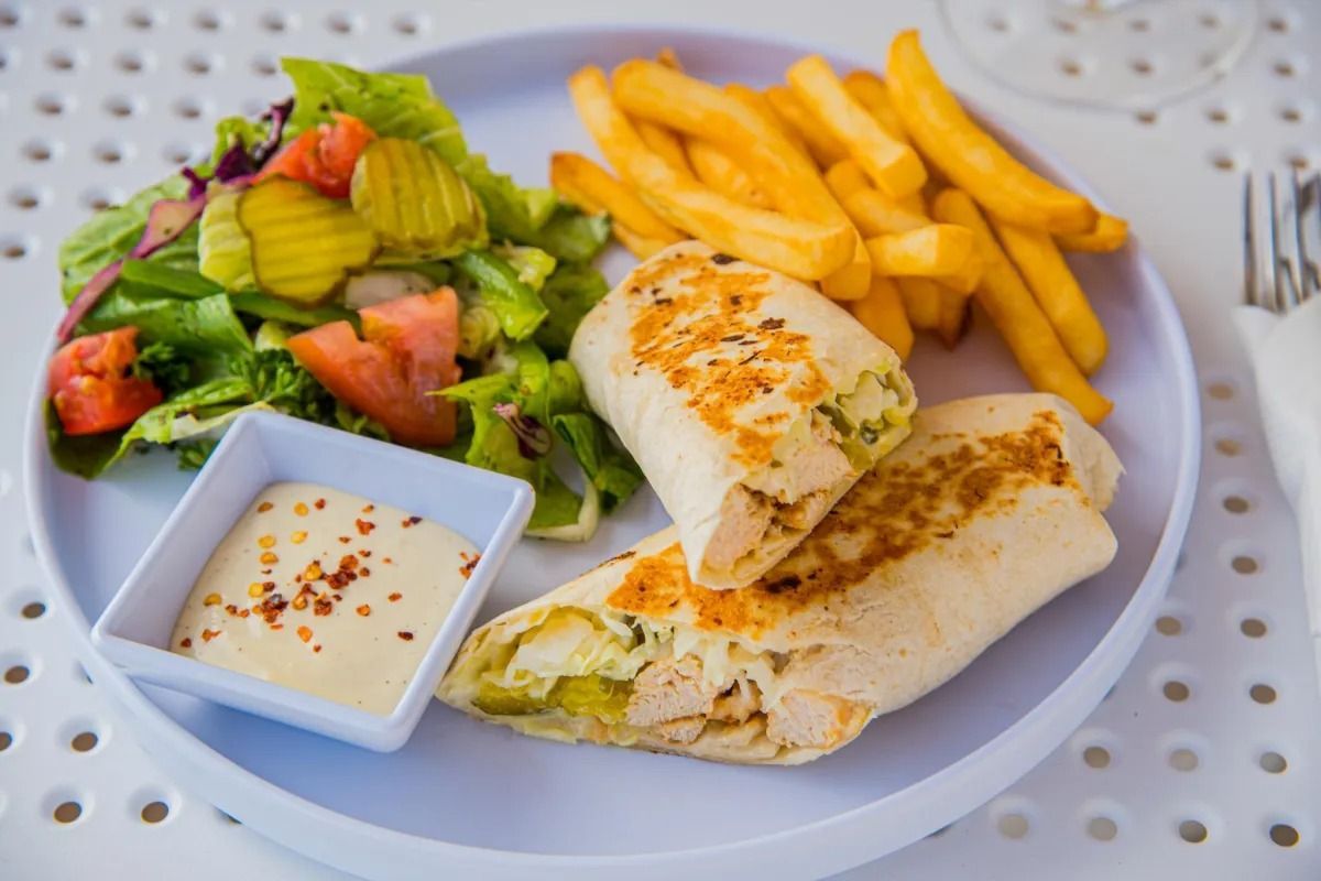 A plate featuring two chicken wraps, a side of french fries, a green salad with pickles and tomatoes, and a dipping sauce.