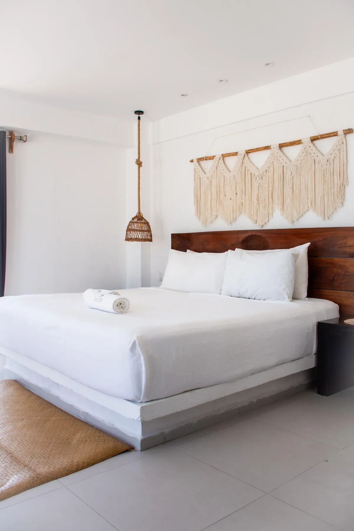 Modern minimalist bedroom with a white bed, wood headboard, macramé wall hanging, and a pendant light.
