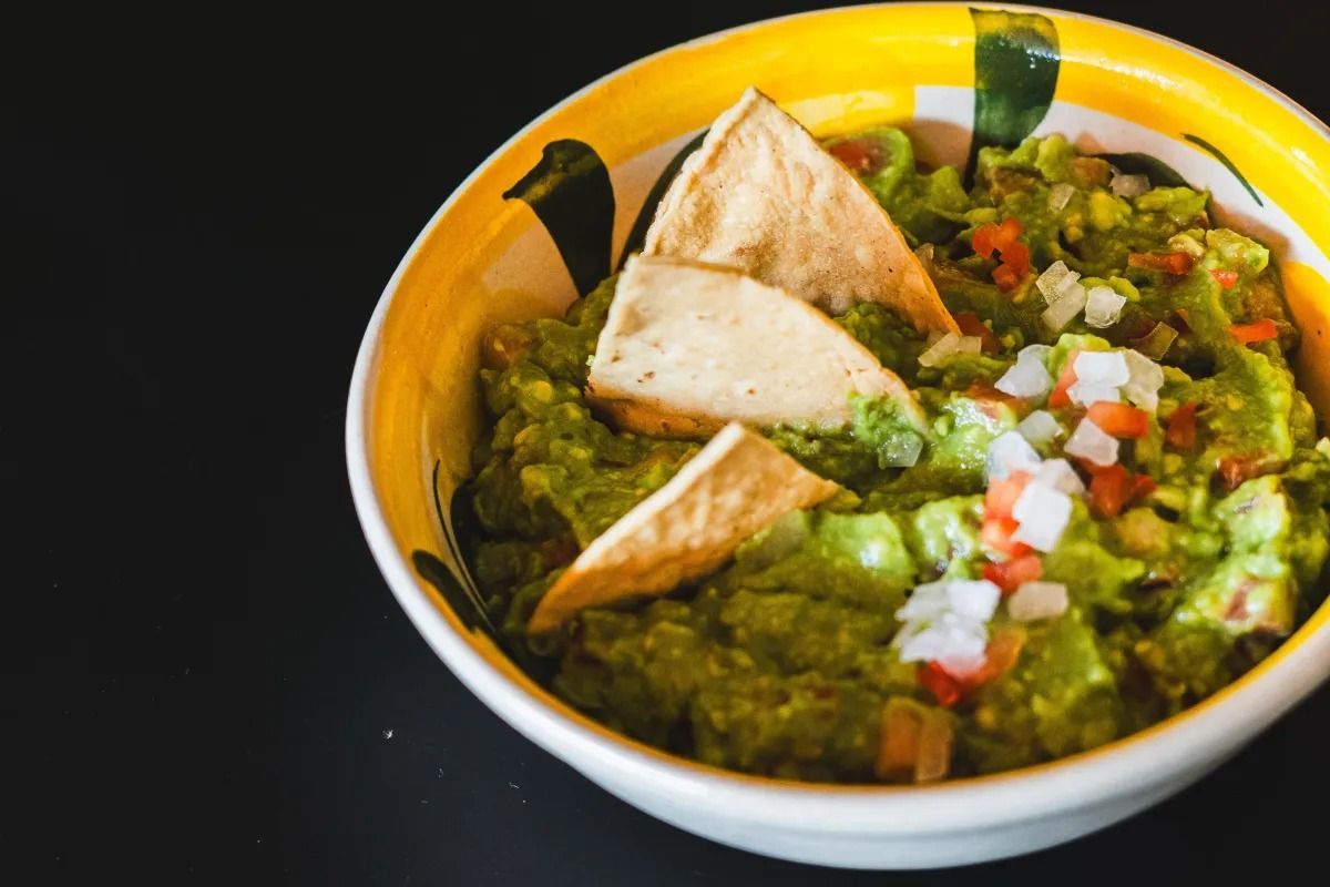 A bowl of chunky guacamole topped with three tortilla chips, diced white onion, and red tomato, set on a black surface.