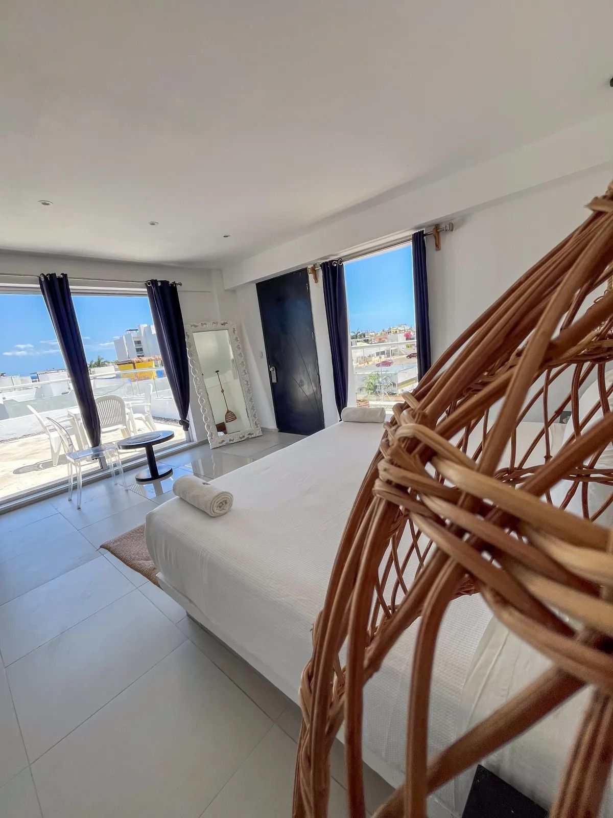 A bright bedroom with a white bed, a large wicker hanging chair in the foreground, and a view of the sea through windows.