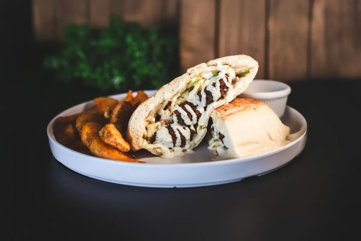 A plate with a halved pita sandwich, potato wedges, and a small cup of sauce, set against a wooden background.