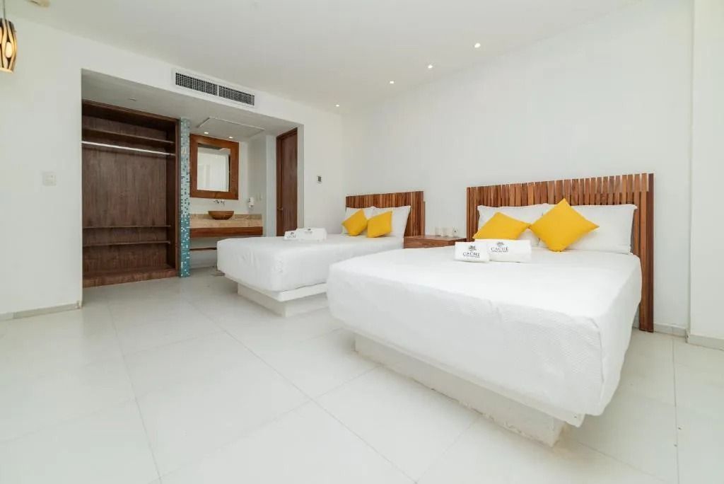 A bright, modern hotel room with two white queen beds, wooden headboards, and an open bathroom area with a vanity.