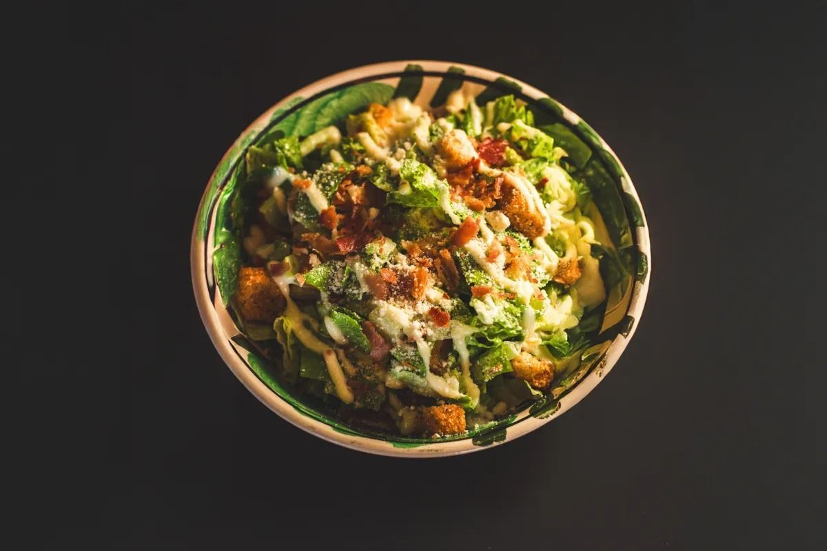 A Caesar salad with croutons, bacon bits, and dressing in a patterned bowl against a dark background.