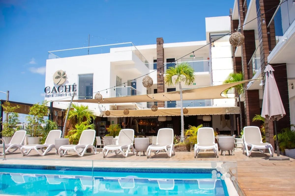 A sunny swimming pool area at the Cachi Boutique Hotel, featuring rows of white lounge chairs under a tan canopy shade.