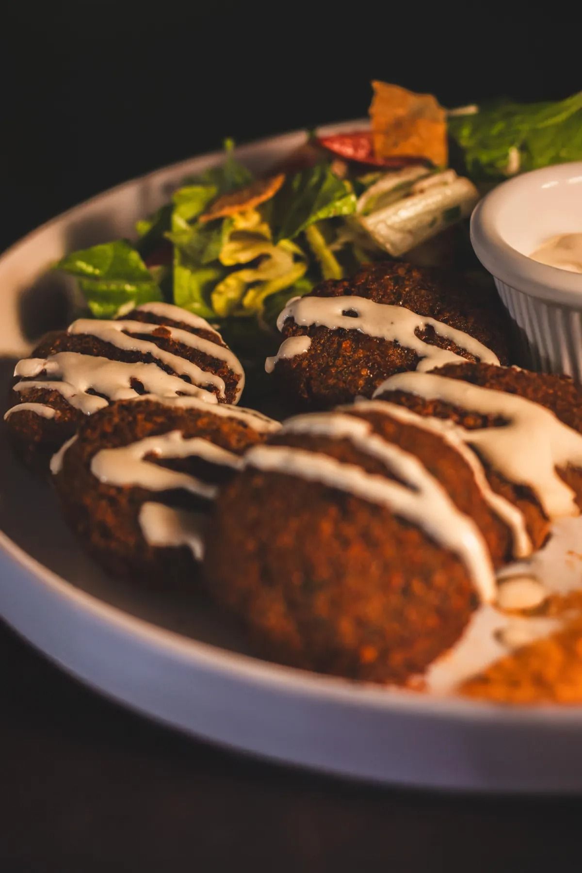 A plate of falafel drizzled with white sauce, served with a side of mixed green salad and a small bowl of dip.