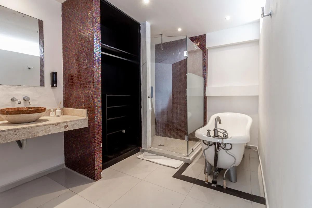 A modern bathroom with a stone sink, an open wardrobe, a glass shower, and a white freestanding bathtub on tiled floors.
