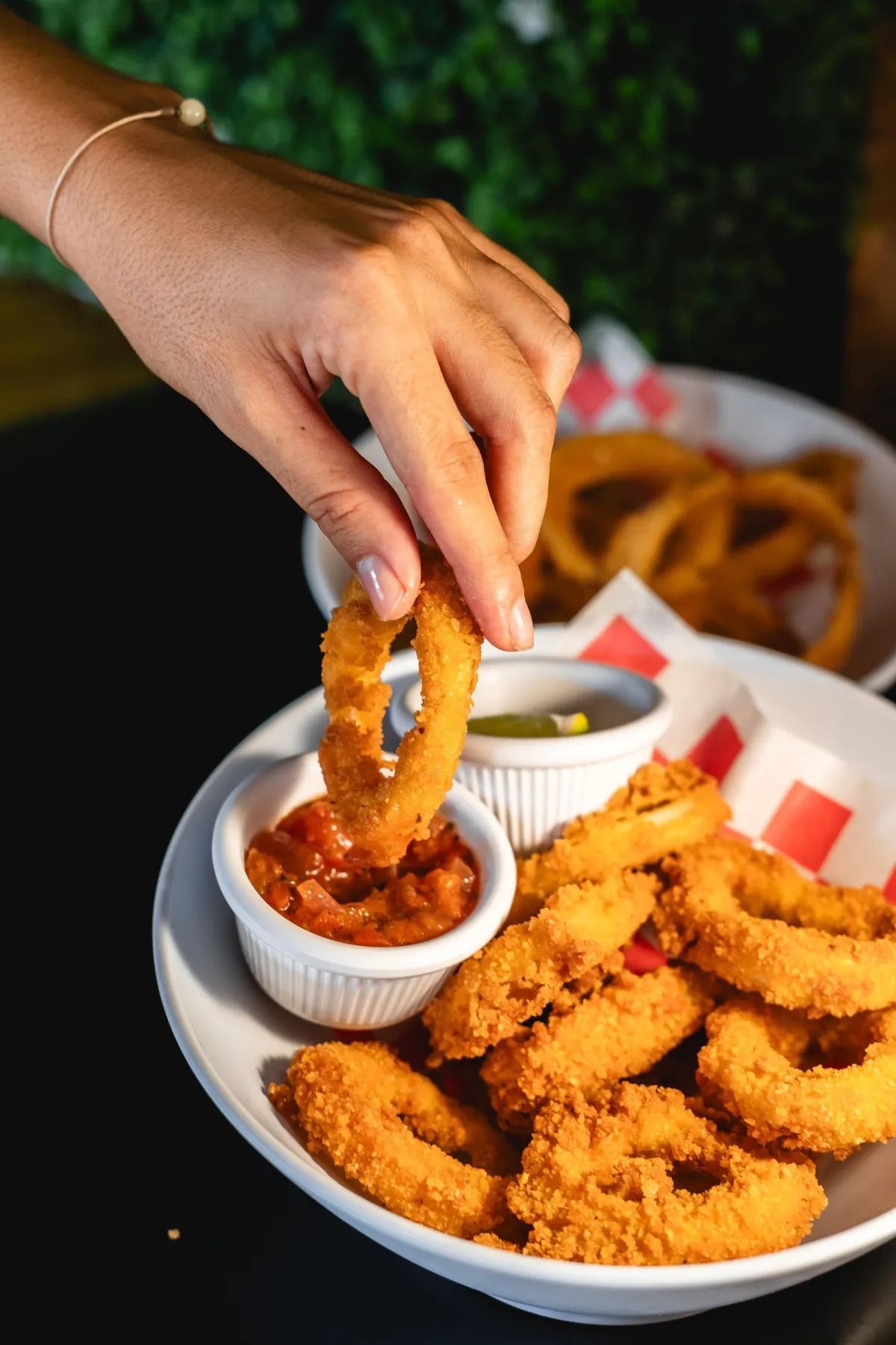 A hand dips a crispy golden onion ring into a small bowl of red sauce on a plate of fried appetizers.
