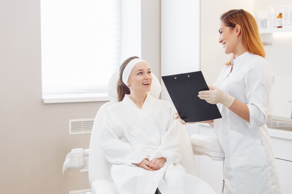 Woman in a spa gown smiles, talking to a doctor who holds a clipboard, both in a well-lit treatment room.