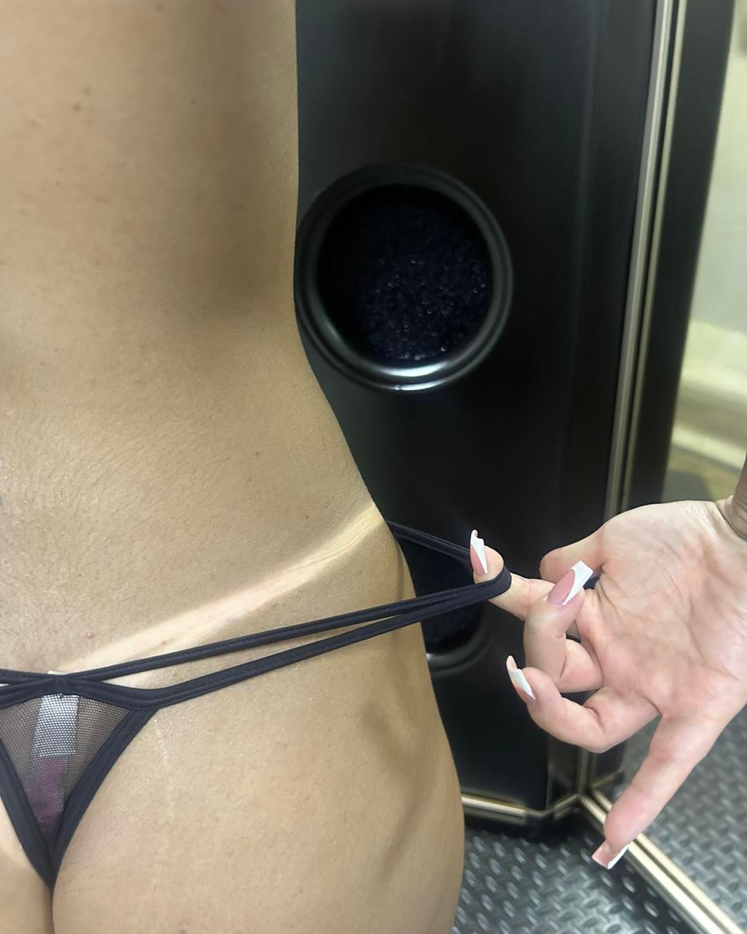 Person getting a spray tan, tan line visible. A hand holds the thin thong. Dark spray tan booth.
