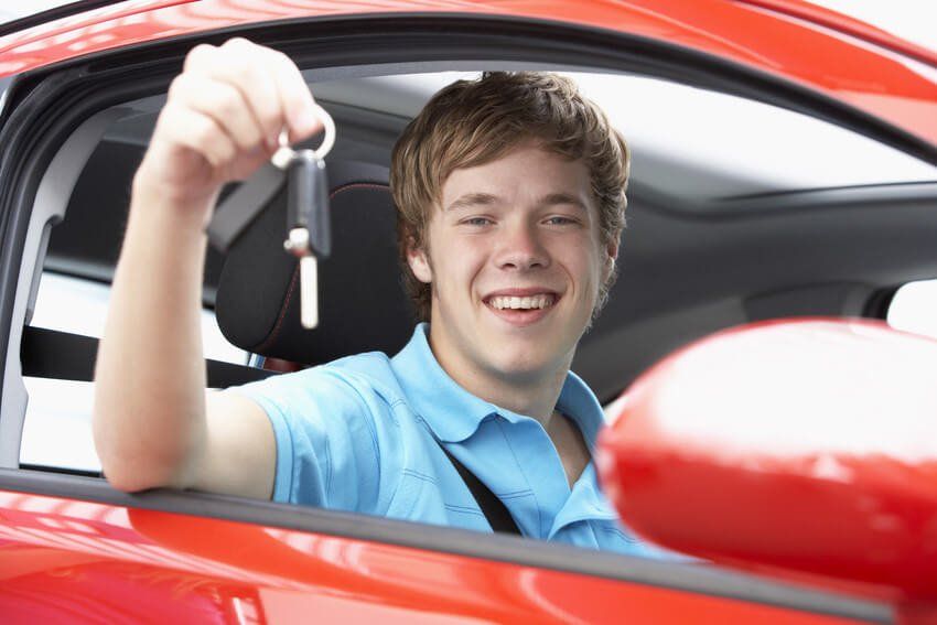 A young man in a blue shirt, holding up a set of car keys. He is sat in a red car.
