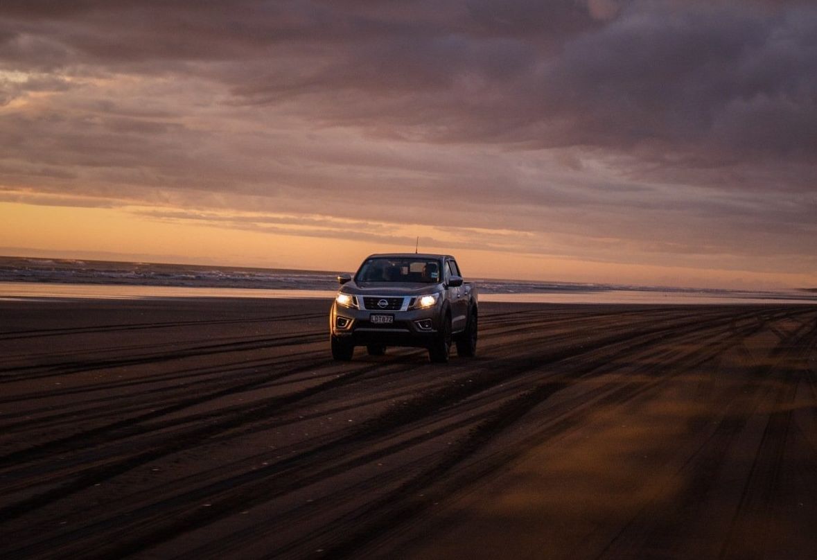 Dark pickup truck driving on a beach at sunset; orange and purple sky.