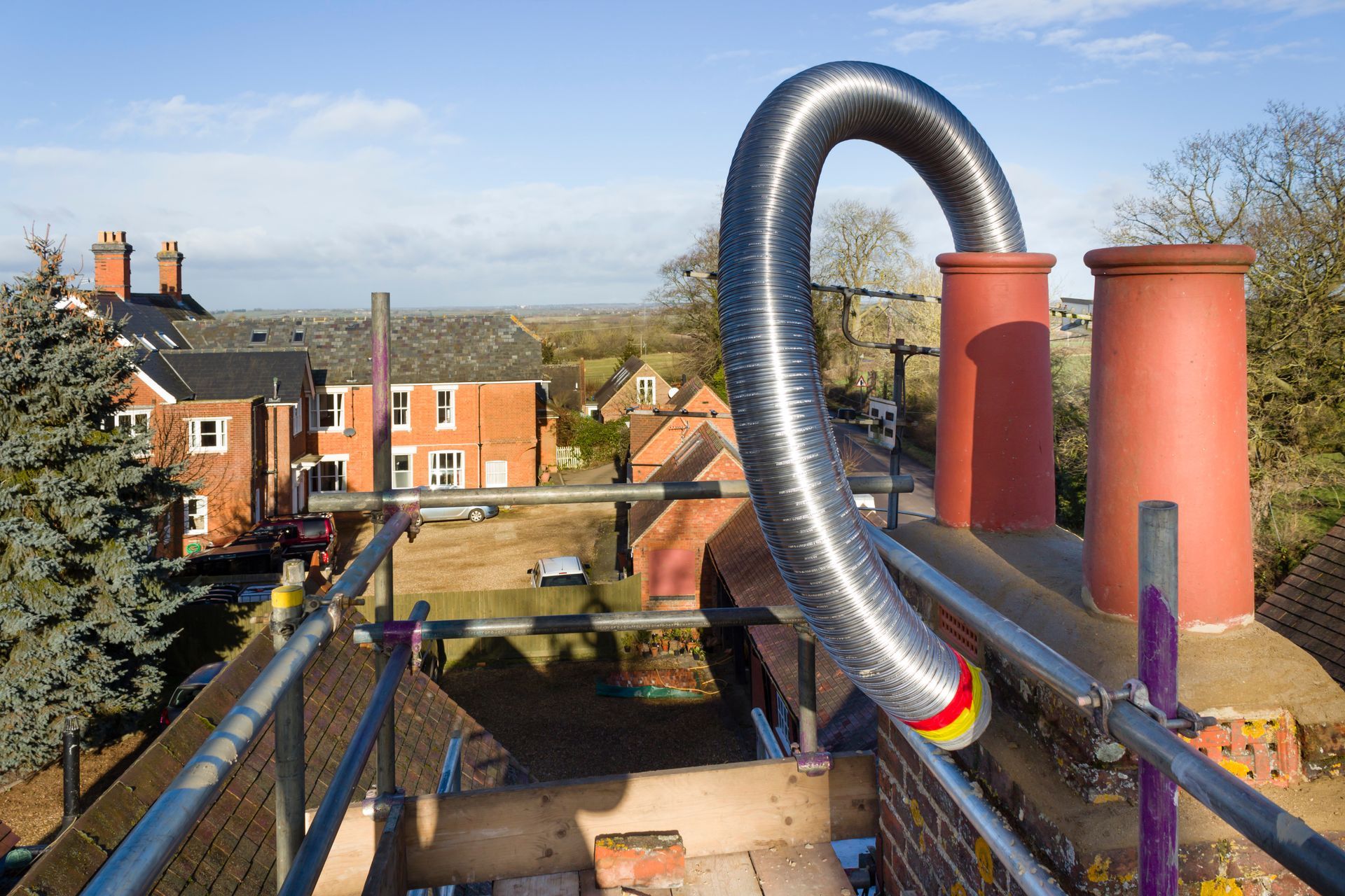 A metal flexible flue liner loops out of a brick chimney on a roof with scaffolding, set against a sunny rural landscape. A metal flexible flue liner loops out of a brick chimney on a roof with scaffolding, set against a sunny rural landscape.
