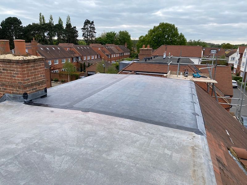 A partially covered flat roof under construction, featuring dark roofing material, light concrete sections, and a chimney.