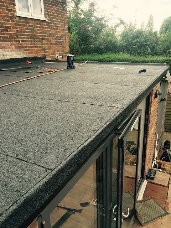 A flat roof covered in dark, textured felt material, adjacent to a brick wall with windows above a doorway.