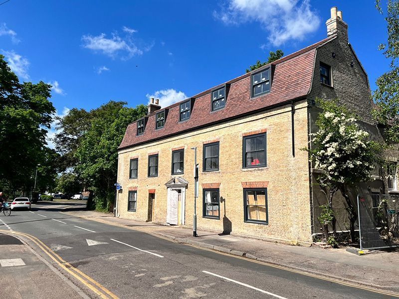 A three-story yellow brick building with a reddish tiled roof and dormer windows, situated along a street under a blue sky.