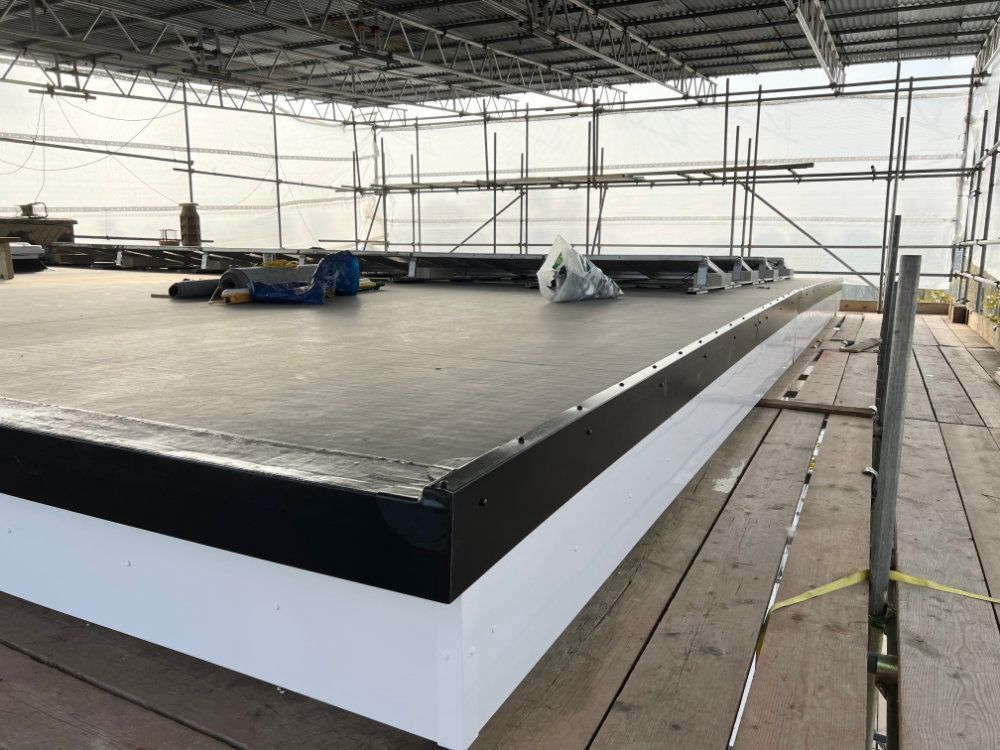 A flat roof under construction, featuring black membrane surfacing and white trim, surrounded by temporary scaffolding.