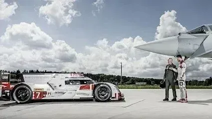 Window Tinting Service-Race car beside a fighter jet on a tarmac. Two men stand talking, under the jet's wing, on a cloudy day.