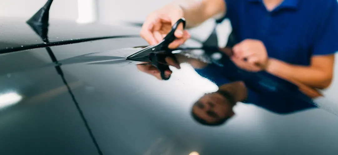 A man is cutting a piece of tinted glass on a car with scissors.