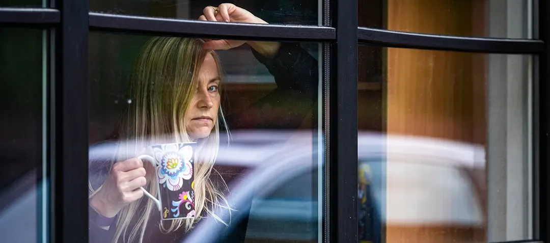 A woman is looking out of a window while holding a cup of coffee.