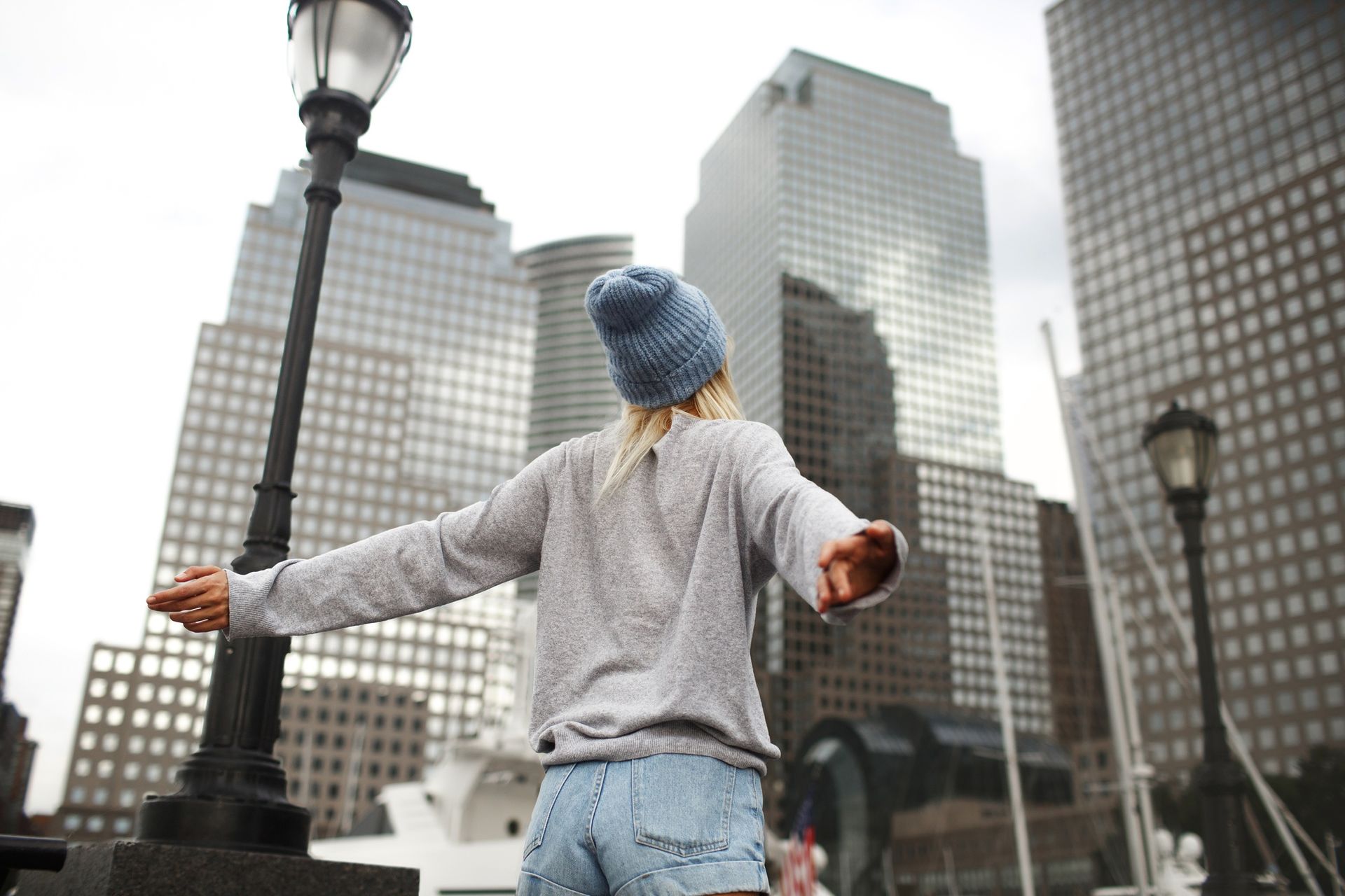 Person in blue hat stands with arms outstretched in front of tall city buildings.