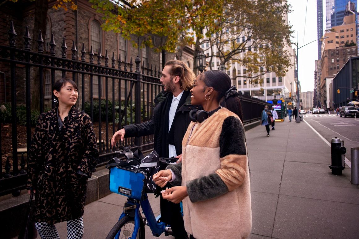 Three people talking beside a blue rental bike on a city sidewalk near a fenced park