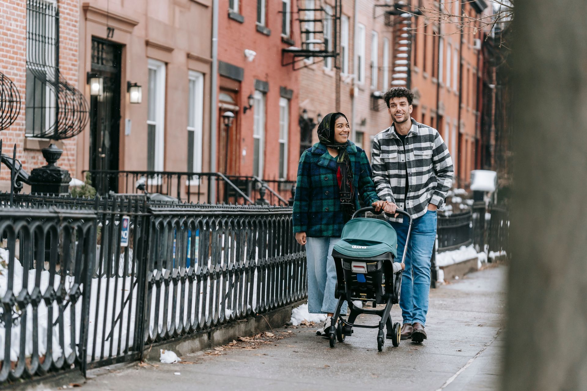Couple walking with a stroller on a wet city sidewalk beside brownstone buildings and iron railings