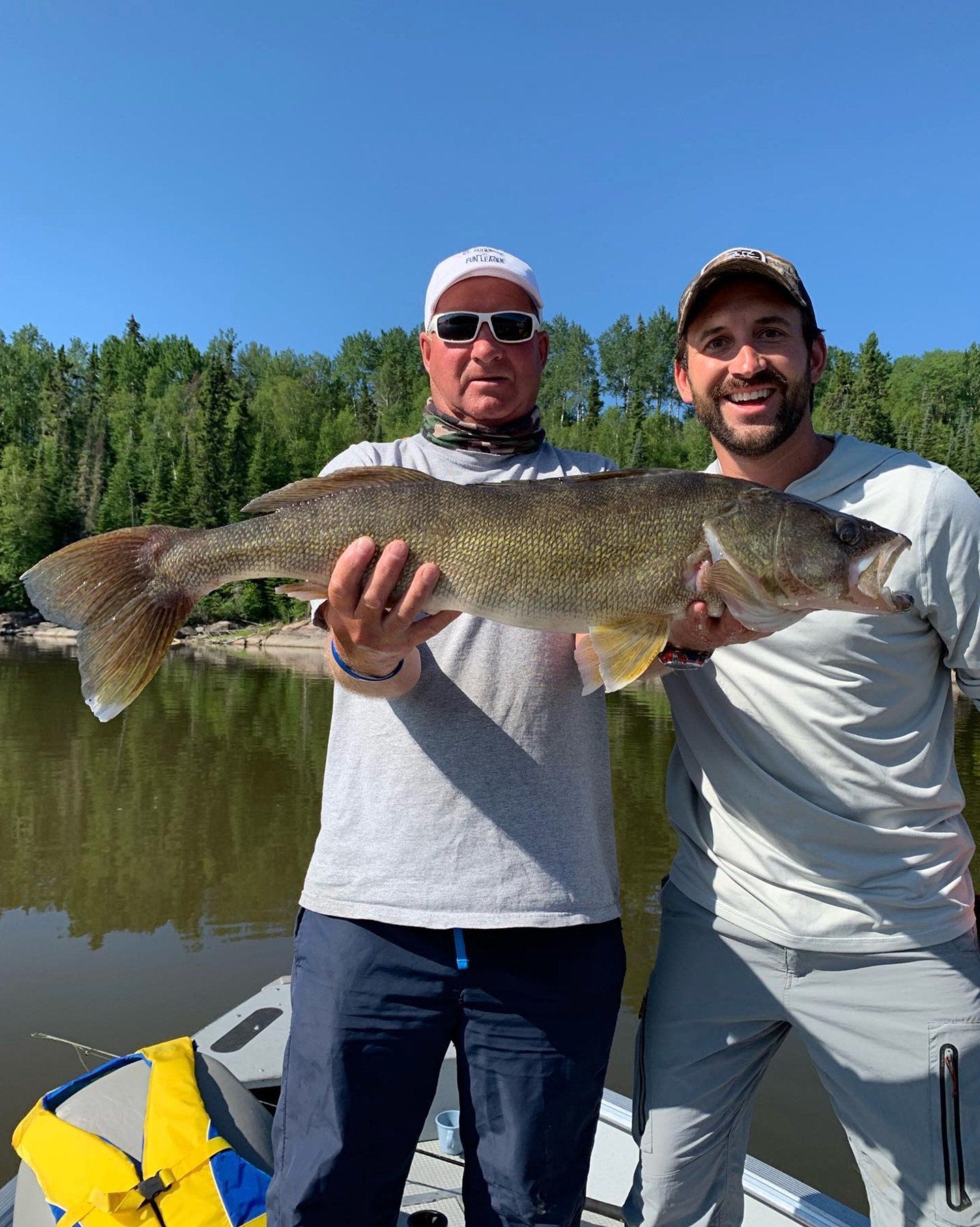 Two men are standing beside each other on a boat holding a large walleye fish.