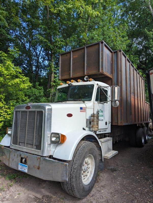 A white garbage truck is parked in front of a forest.