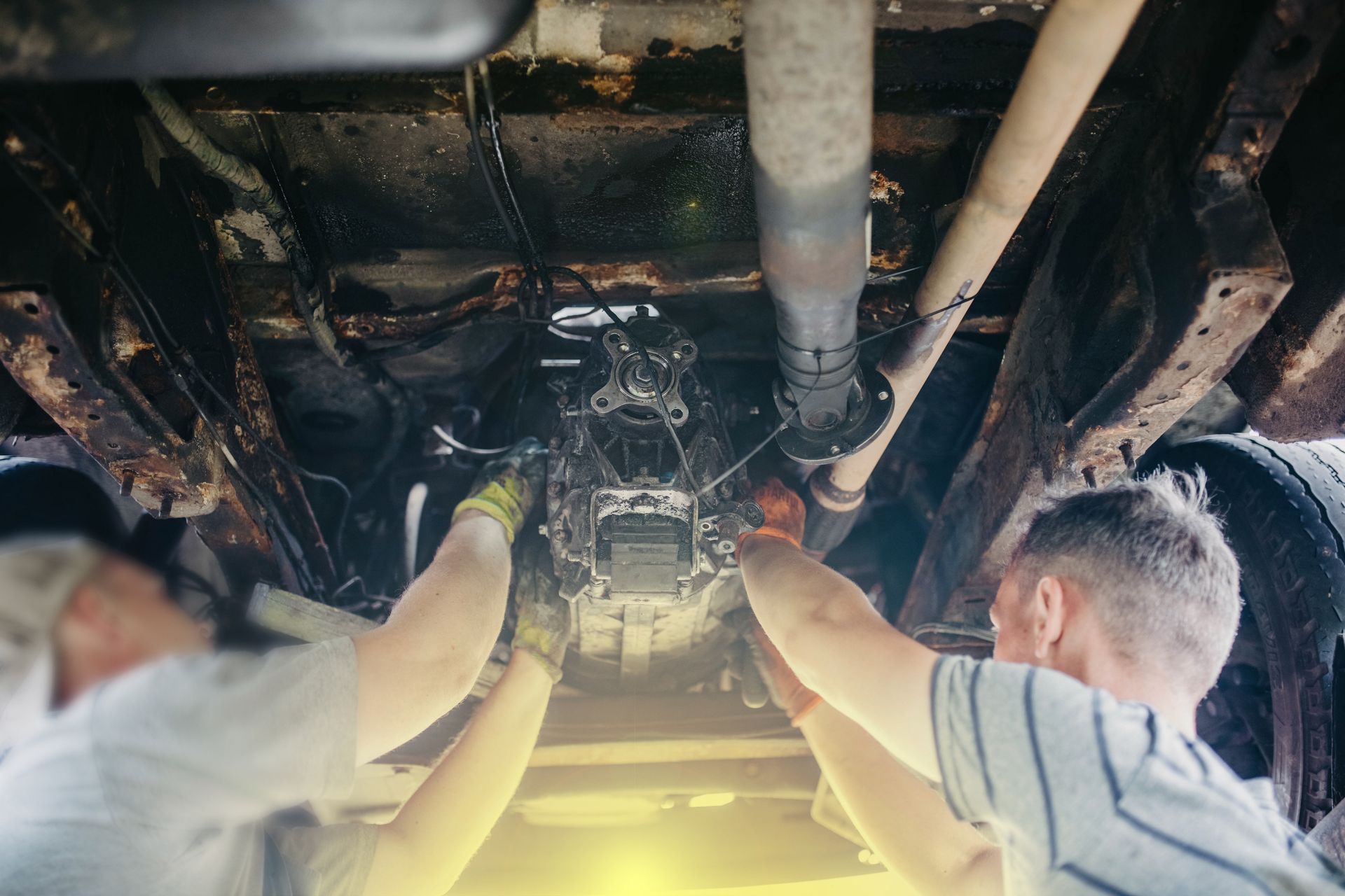 Two people working on the underside of a vehicle. They are using tools on the engine or transmission.