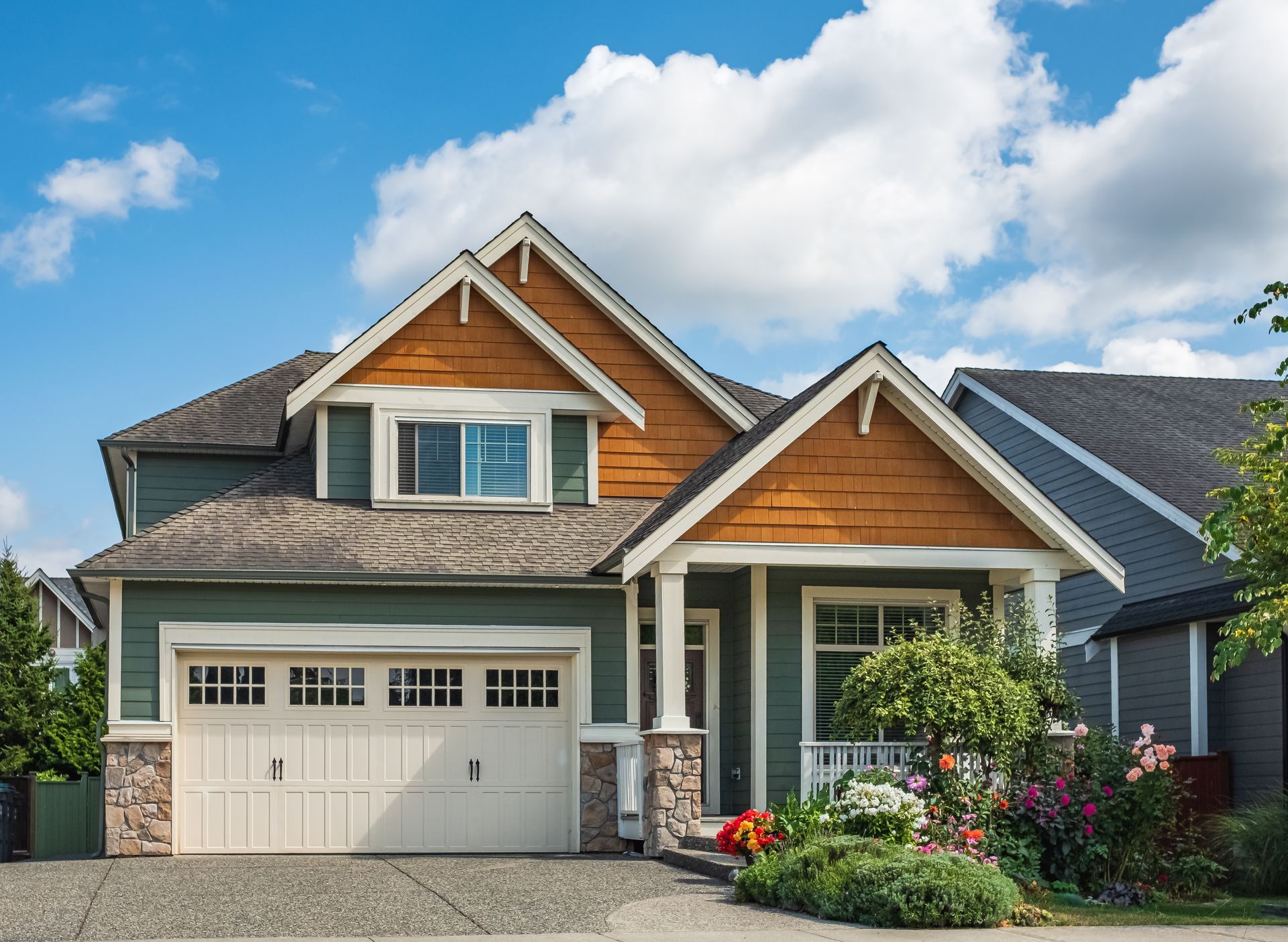 Green two-story house with tan garage door, brown roof accents, and lush front garden against a blue sky.