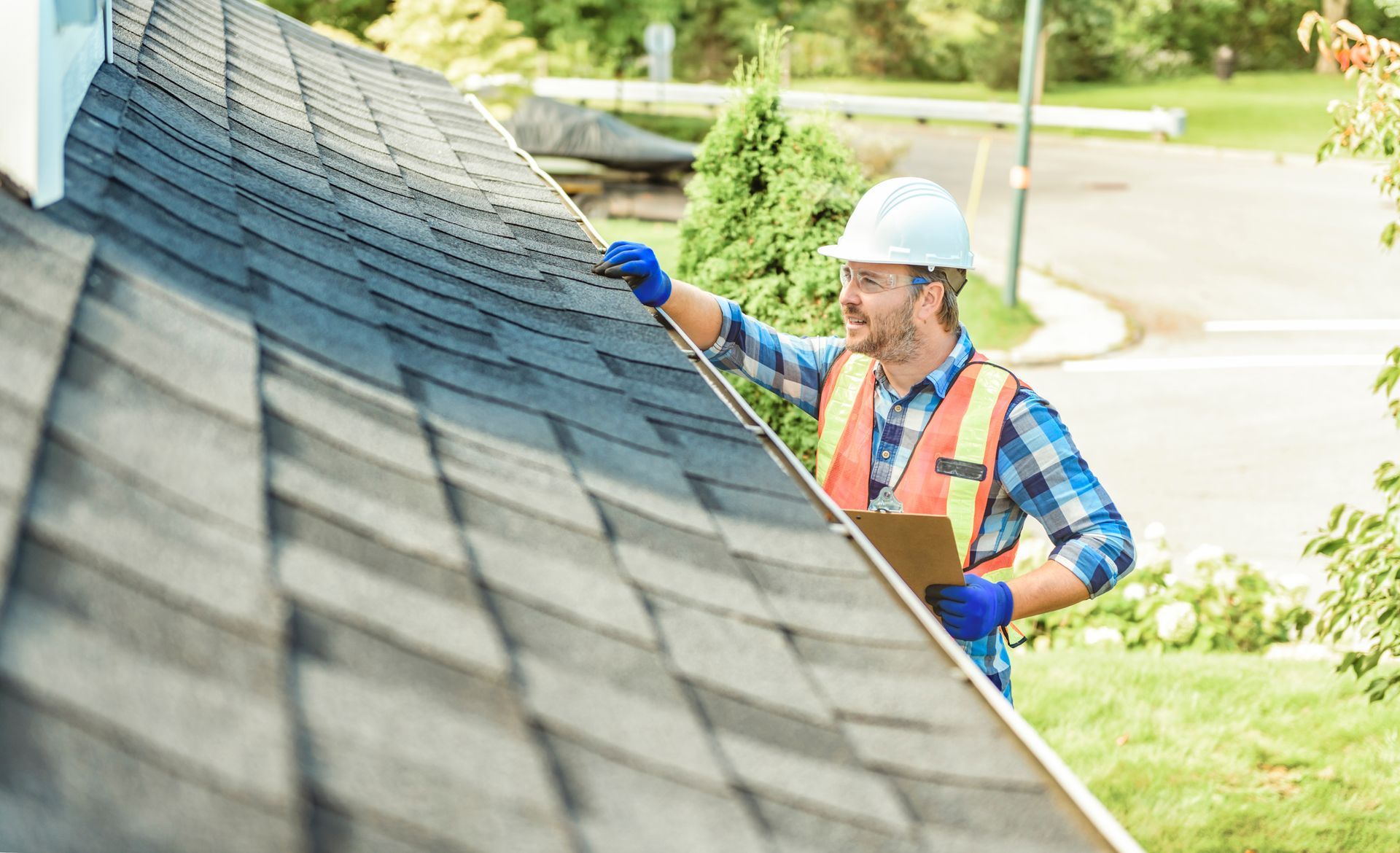 Roofer in hard hat inspecting a dark shingled roof, outdoors on a sunny day.