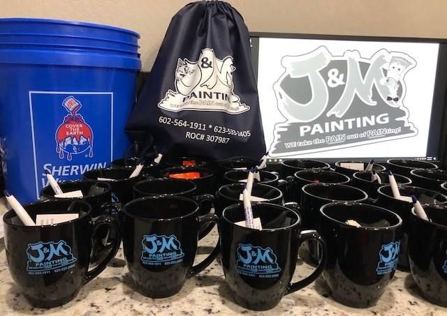 J&M Painting promotional setup: black mugs, blue bucket, drawstring bag, and logo signage.