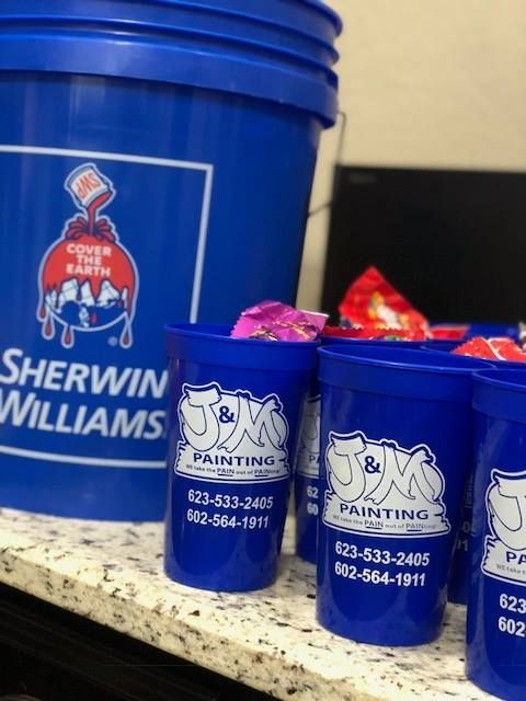 Blue Sherwin Williams paint bucket and J&M Painting cups with candy on a countertop.