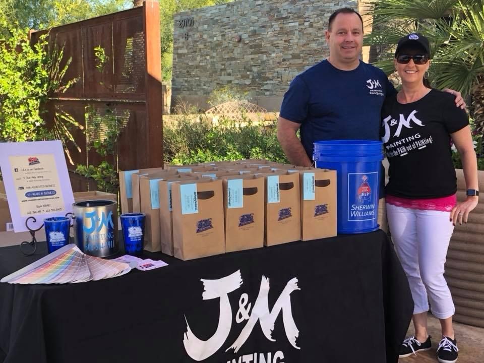 Two people at a J&M Painting display table with brown bags, paint supplies, and a logo.