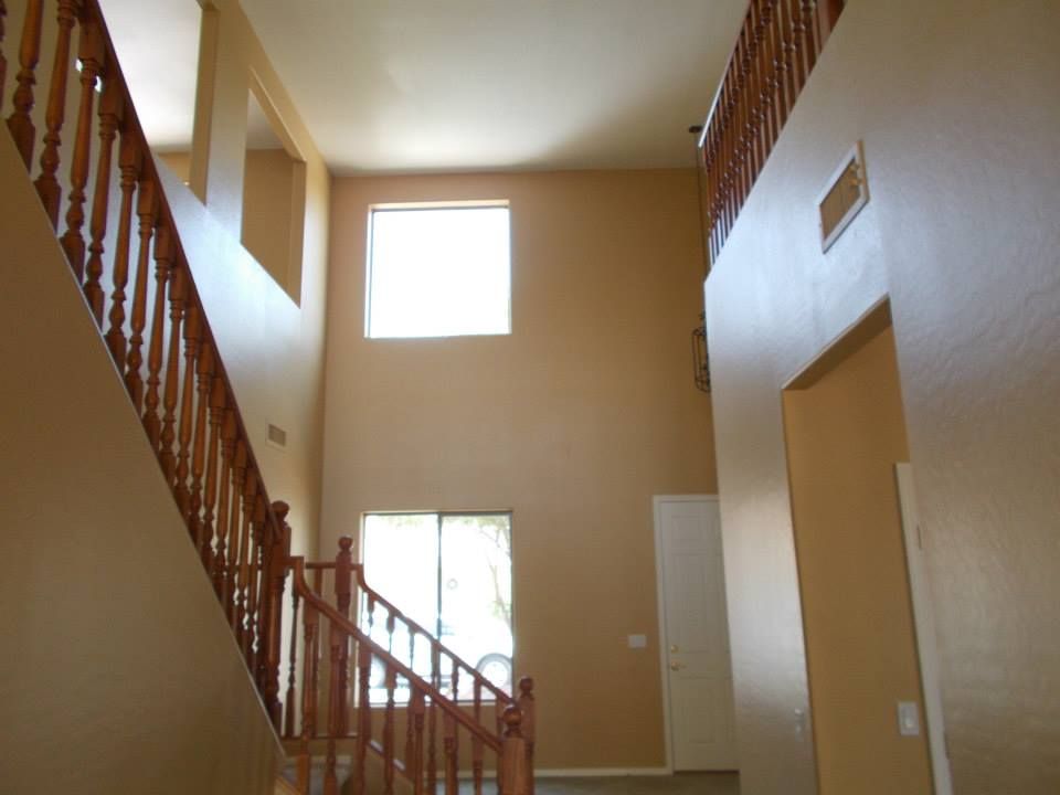 Two-story foyer with staircase, natural wood railing, and beige walls. Sunlight streams through windows.