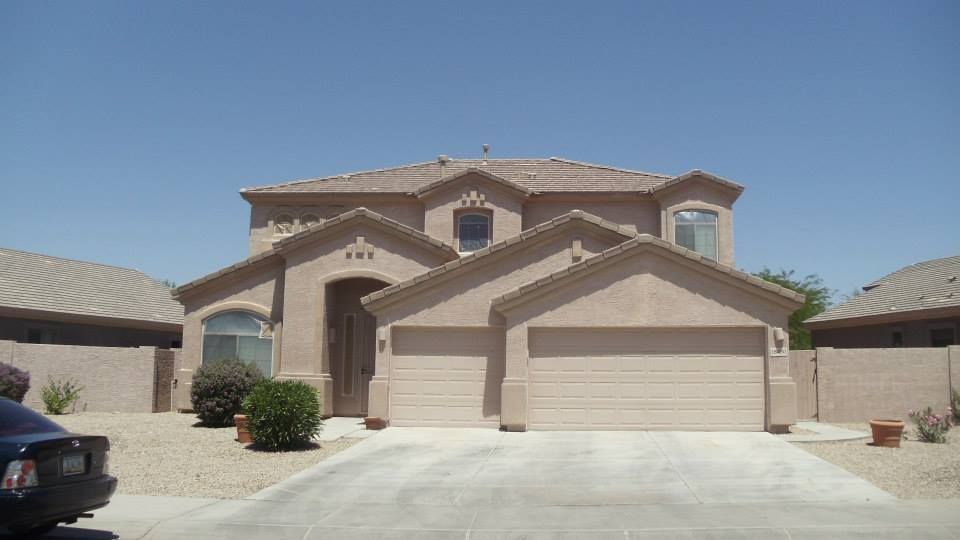 Beige two-story house with a two-car garage, driveway, and dry landscaping under a clear, blue sky.
