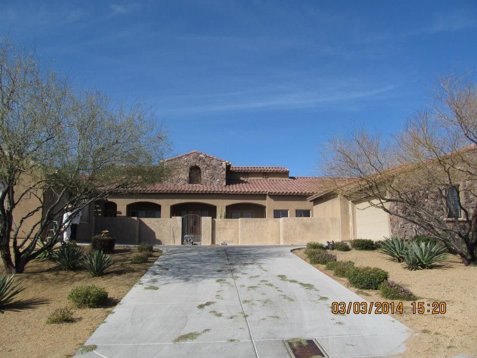 A tan, single-story house with a clay tile roof and a long concrete driveway under a blue sky.