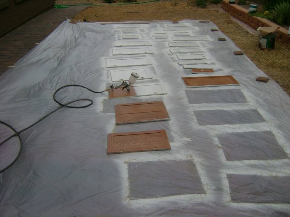 Cabinet doors laid out on a tarp, being painted with a spray gun outdoors.