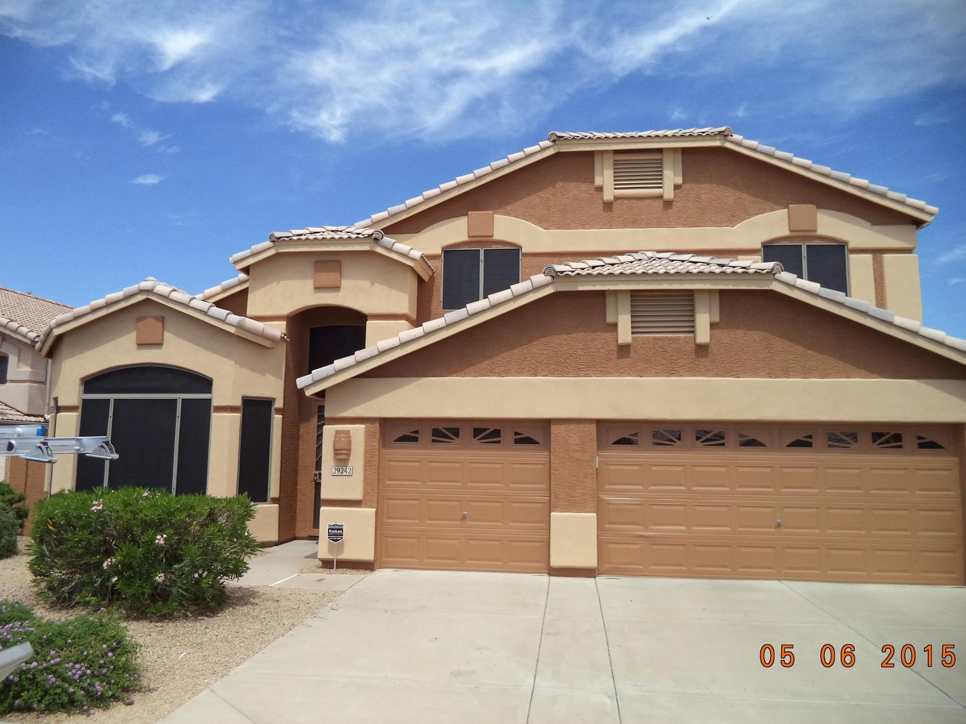 Two-story beige stucco house with tan trim, garage doors, and blue sky.