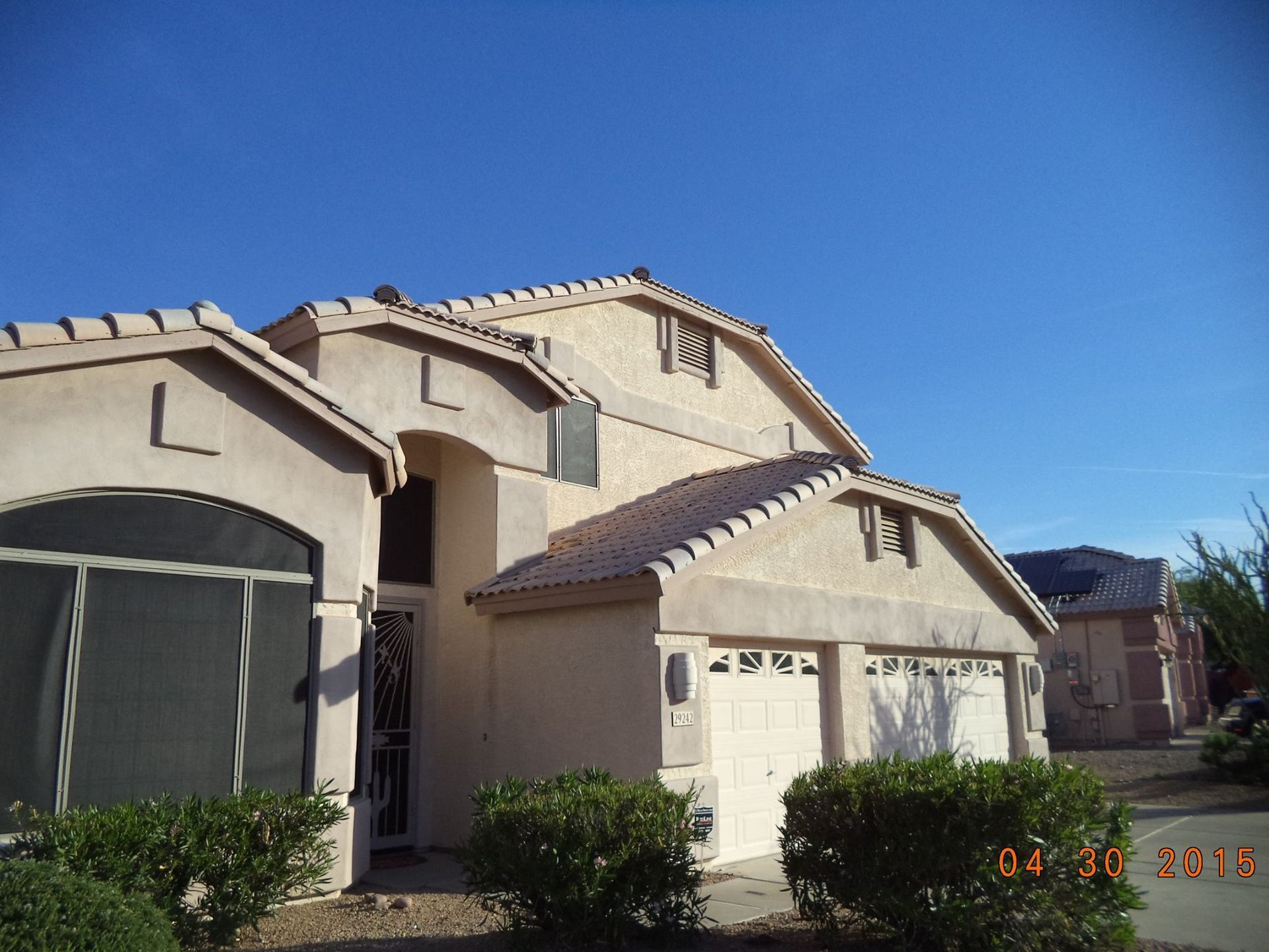 Beige two-story house with attached garage under a clear blue sky; bushes in the foreground.