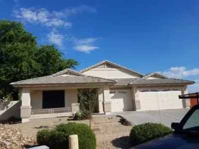 Beige stucco house with a two-car garage under a blue sky. Bushes and a tree are in front.