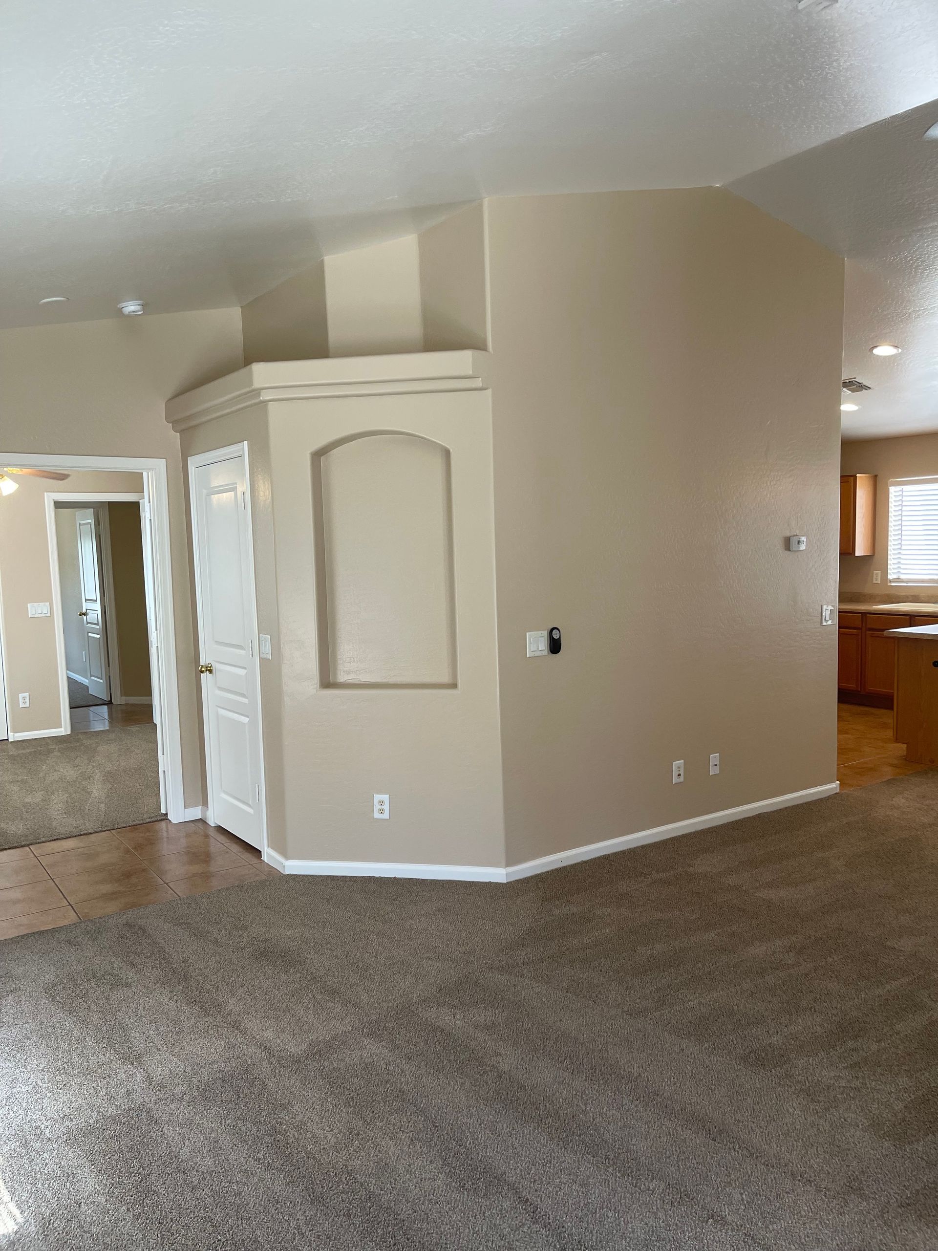Beige-walled living room with a door, arched niche, and kitchen in the background. Brown carpet.