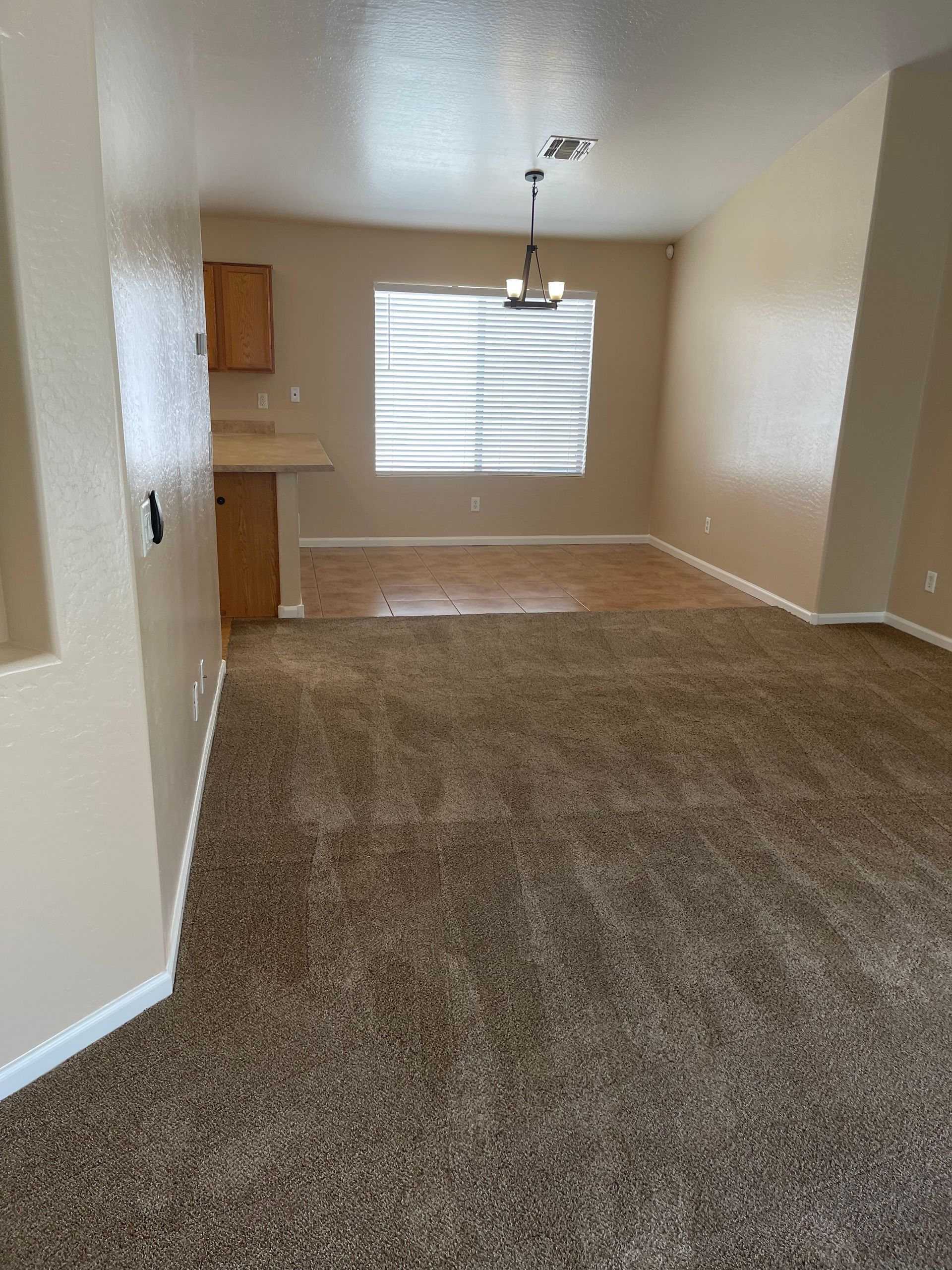 Beige living room with brown carpet, leading to a dining area with window and wood flooring.