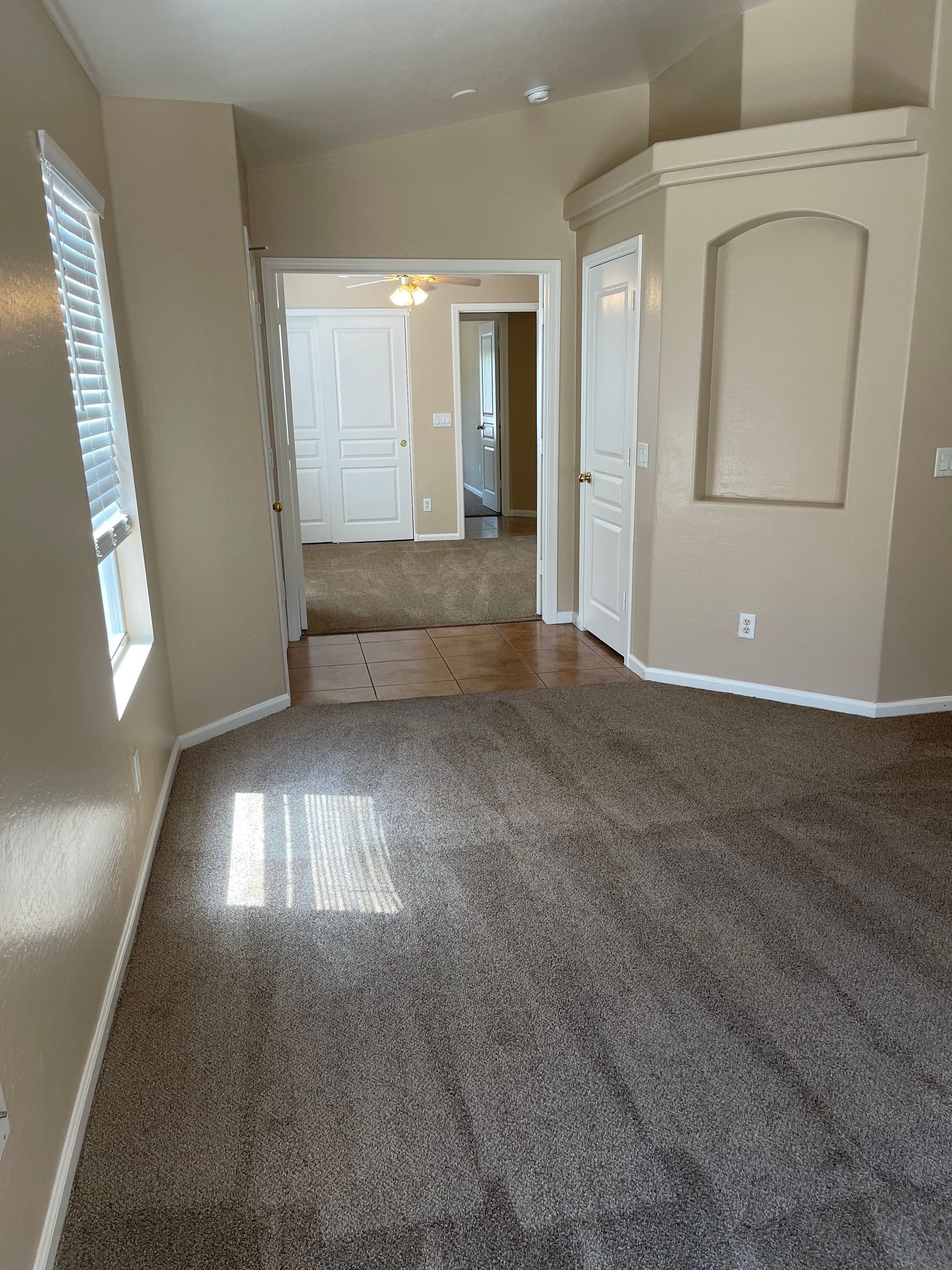 A beige-walled living room with carpet, doorway to a hallway and bedroom, and a window with blinds.