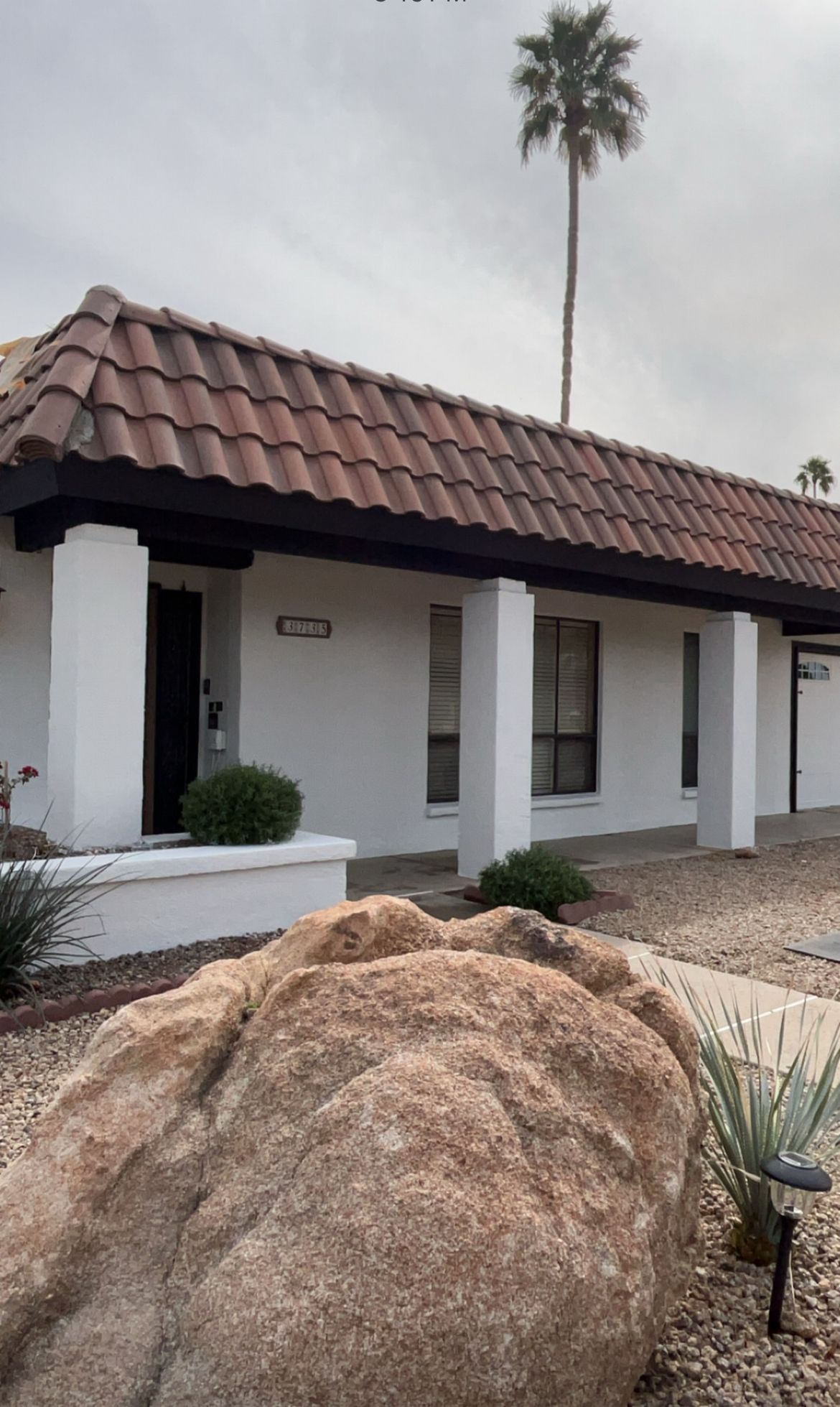 White stucco house with red tile roof, large rock in foreground, tall palm tree in background.